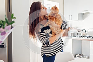 Young woman playing with cat in kitchen at home. Girl holding and hugging ginger cat