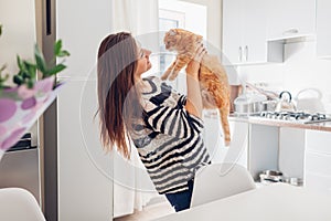 Young woman playing with cat in kitchen at home. Girl holding and raising red cat