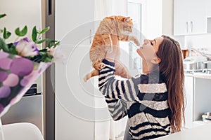 Young woman playing with cat in kitchen at home. Girl holding and red raising cat