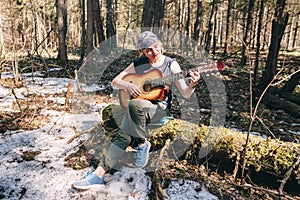 Young woman playing guitar sitting on old tree covered with moss at sunny spring forest