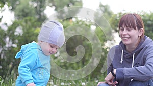 Young woman is playing with her little baby son in the city park in a summer cloudy day by blowing dandelion with trees