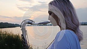 Young woman playing on the saxophone near the riverbank at dawn, a beautiful girl in a long dress with a wind musi