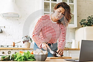 Young woman plus size body positive in red longsleeve cooking food using laptop with online video recipes on kitchen at home