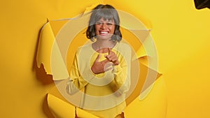 Young woman points at camera giggles happily, breaks through paper background