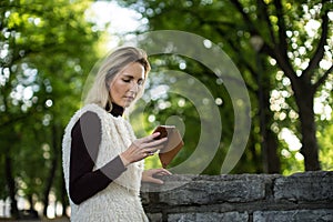 Young woman portrait in summer. Blonde girl is reading message on cell phone outside in city nature. Female with telephone.
