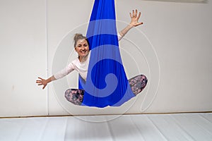 A young woman poses while doing anti-gravity aerial yoga in a blue hammock on a white background.