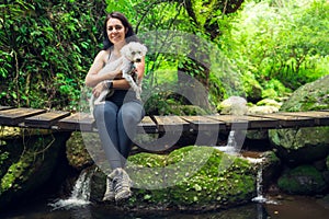 Young woman posing with her puppy on suspension bridge in tropical forest. Woman and pet in green wet forest on sunny day.