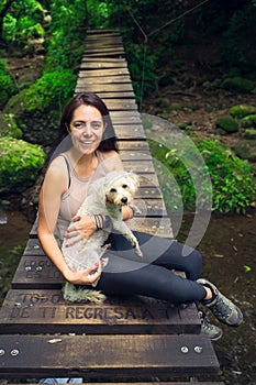 Young woman posing with her puppy on suspension bridge in tropical forest. Woman and pet in green wet forest on sunny day.
