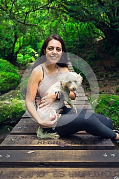 Young woman posing with her puppy on suspension bridge in tropical forest. Woman and pet in green wet forest on sunny day.