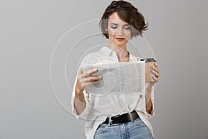 Young woman posing isolated over grey background drinking coffee holding newspaper reading