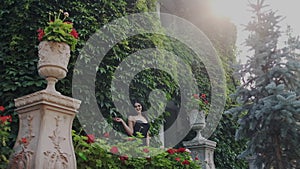 Young woman posing in a luxurious large garden or homestead. She stands against a green wall of grapes and touch flowers