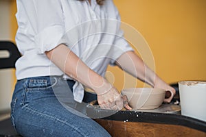 Young woman potter in apron at work. Craftsman artist shapes the jug with her hands and a special tool on pottery wheel.