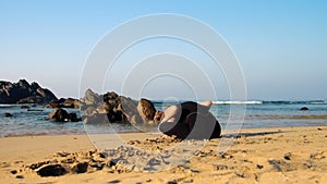 Young woman practices yoga on beach against brown rocks