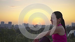 A young woman practices yoga on a mountain in the background of a big city