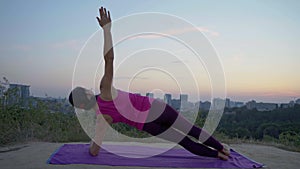 A young woman practices yoga on a mountain in the background of a big city