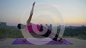 A young woman practices yoga on a mountain in the background of a big city