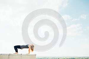 Young woman practices yoga outside. Blonde girl do purvottanasana or upside down pose on roof. She dressed in black