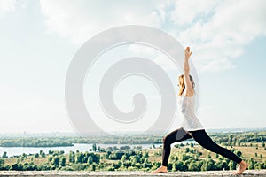 Young woman practices yoga outside. Blonde girl standing in Warrior I posture Virabhadrasana 1 , full length on parapet