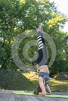 Young woman practices yoga in the pose of Shirshasana. Athletic woman stands on her head. Morning yoga in park