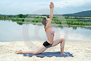 Young woman practices yoga at sand beach near lake with mountain background