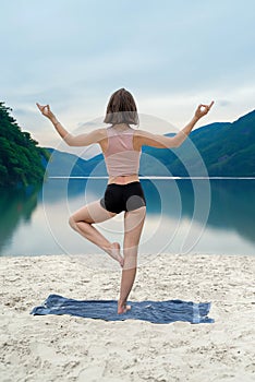 Young woman practices yoga at sand beach near lake with mountain background