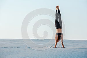 Young woman practicing inversion balancing yoga pose handstand on sand.