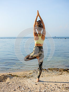 Young woman practicing Vrikshasana, Tree Pose on the beach. Hands in namaste mudra. Yoga retreat. Healthcare concept. View from