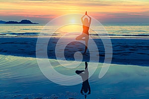 Young woman practicing yoga on the beach at sunset with reflection in water.