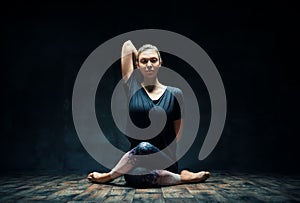 Young woman practicing yoga doing Gomukhasana, cow face pose, in dark room