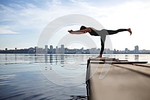 Young woman practicing yoga exercise at quiet wooden pier with city background. Sport and recreation in city rush