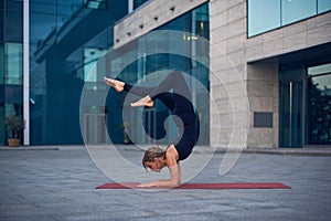 Young woman practicing yoga, standing in Vrischikasana Scorpion pose outdoors against the background of a modern city