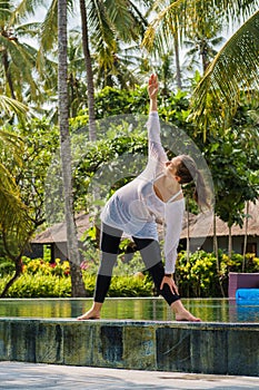 Young woman is practicing yoga in stretching position on the swimming pool with green plants, palm trees around in hotel