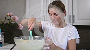 Young woman prepares dough mixing ingredients in the the bowl using whisk in the kitchen. Homemade food. Slowmotion shot