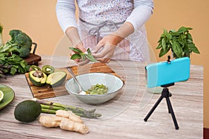Young woman preparing detox juice while making video lesson online with phone - Girl making smoothie with green vegetables and
