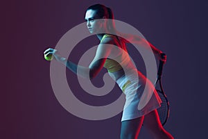 Young woman, professional tennis player training isolated on blue background in neon.