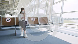 Young woman in a protective face mask sitting in the airport lounge and uses her smartphone. Sends text messages, looks