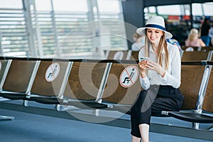 Young woman in a protective face mask sitting in the airport lounge and uses her smartphone. Sends text messages, looks