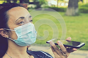 Young woman in protective mask holding a smartphone. Woman siting on bench in green park with mobile phone. First stage of