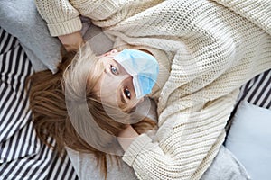 Young woman in protective mask lying on bed, top view