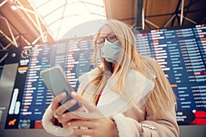 Young woman in protective mask with phone and backpack in front of information board in airport terminal. Concept