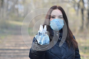 Young woman in protective mask shows sanitizer spray bottles outdoors in spring wood