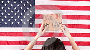 Young woman protester holds cardboard with Bans Of My Body signs against USA flag on background. Girl protesting against anti-