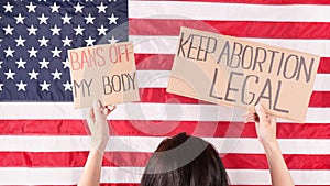 Young woman protester holds cardboard with Keep Abortion Legal and Bans Of My Body signs against USA flag on background. Girl