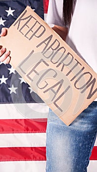 Young woman protester holds cardboard with Keep Abortion Legal sign against USA flag on background. Girl protesting against anti-