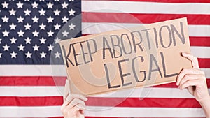 Young woman protester holds cardboard with Keep Abortion Legal sign against USA flag on background. Girl protesting against anti-