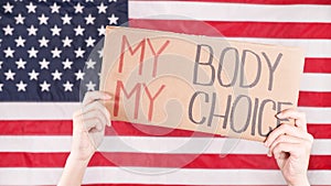 Young woman protester holds cardboard with My Body My Choice sign against USA flag on background. Girl protesting against anti-