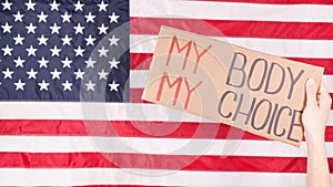 Young woman protester holds cardboard with My Body My Choice sign against USA flag on background. Girl protesting against anti-