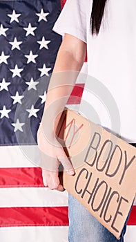 Young woman protester holds cardboard with My Body My Choice sign against USA flag on background. Girl protesting against anti-