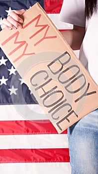 Young woman protester holds cardboard with My Body My Choice sign against USA flag on background. Girl protesting against anti-