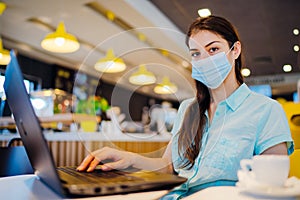 Young woman in a public place working on laptop, wearing protective face mask indoors.Online training education and freelance work